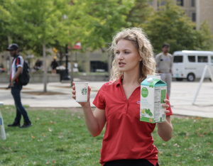 student in red polo with curly blonde hair holds a Starbucks coffee cup and milk cartoon outside, she is speaking to other students